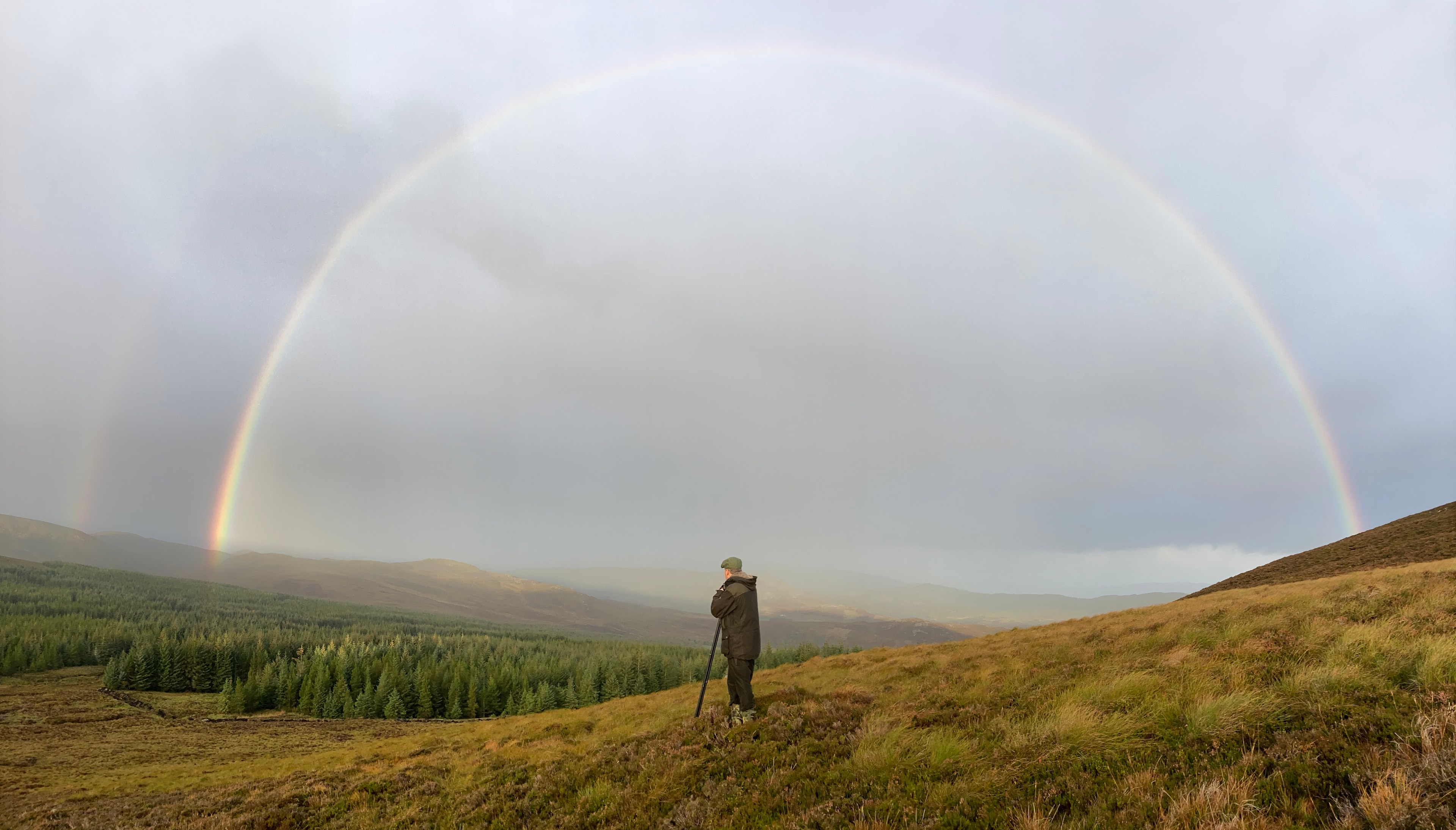 Stalker standing on a hillside beneath a full double rainbow over a Scottish glen